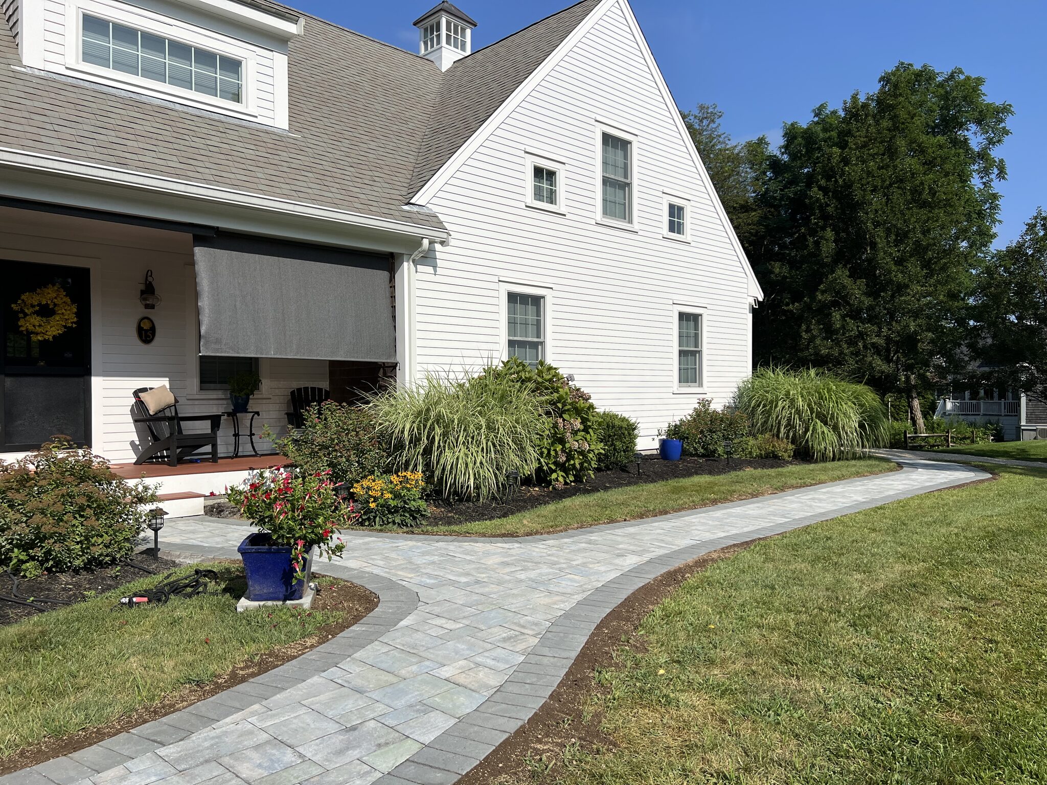 Paver walkway through a landscaped front yard to a white Cape Cod home